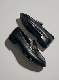 Volterra Black - מוקסין עור שחור עם רצועה רחבה בחזית ועיטור מתכתי זהוב, תפר מודגש סביב החרטום וסוליה דקה.