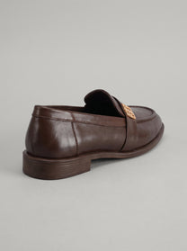 Volterra Dark Brown - מוקסין עור בחום כהה עם רצועה רחבה בחזית ועיטור מתכתי זהוב, תפר מודגש סביב החרטום וסוליה דקה.