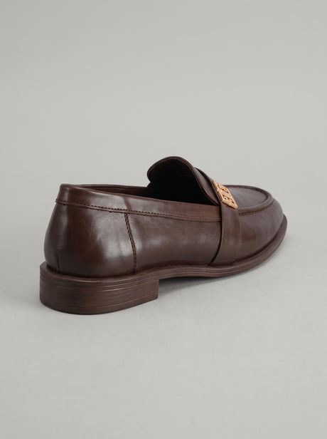Volterra Dark Brown - מוקסין עור בחום כהה עם רצועה רחבה בחזית ועיטור מתכתי זהוב, תפר מודגש סביב החרטום וסוליה דקה.